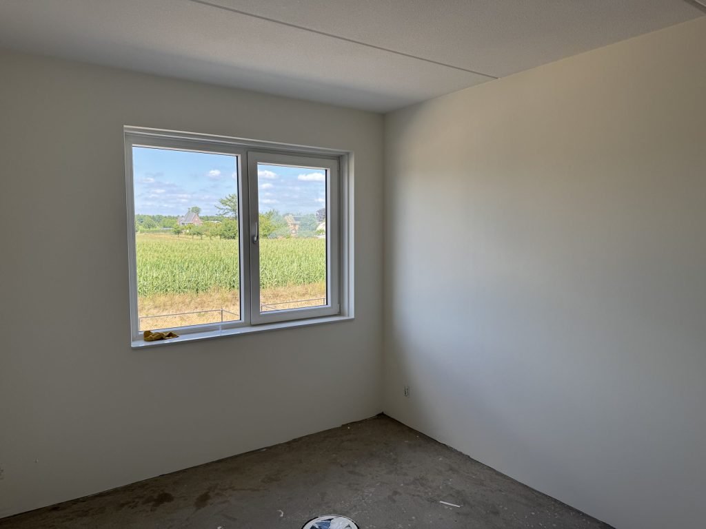 Lichte slaapkamer met gladde witte wanden en een raam dat uitkijkt op een maisveld